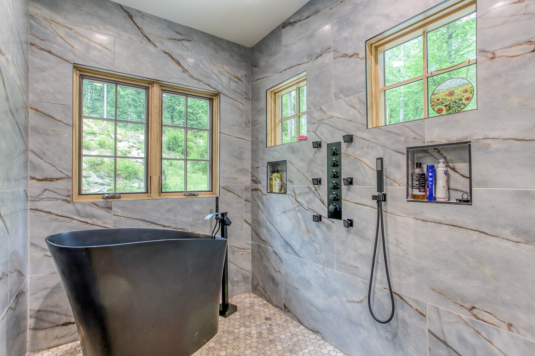 bathroom with all walls covered in grey marble like tile with view of outside forest and standing tub and rainhead shower